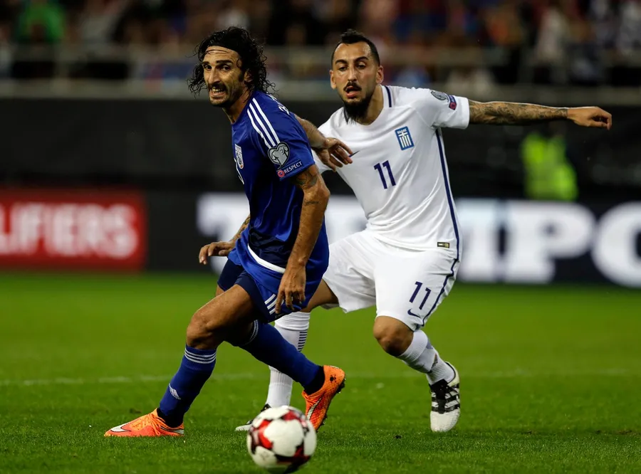 Elias Charalambous, pe vremea când era jucător, într-un duel cu Kostas Mitroglou, în timpul unui meci Grecia - Cipru, disputat în octombrie 2016, în preliminariile pentru Cupa Mondială din 2018. Sursă foto: Profimedia